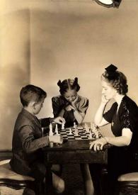 Plano general de Paul Rubinstein (perfil derecho) y Aniela Rubinstein (perfil izquierdo) jugando al ajedrez mientras Eva Rubinstein les observa