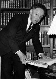 Plano general de Arthur Rubinstein posando inclinado hacia una mesa mientras sujeta en sus manos un libro abierto