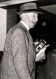 Plano medio de Arthur Rubinstein (perfil derecho) sonriendo, con sombrero y varios regalos en la mano