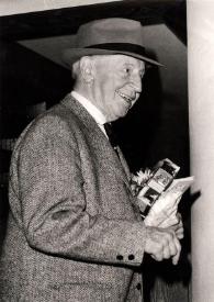 Plano medio de Arthur Rubinstein (perfil derecho) sonriendo, con sombrero y varios regalos en la mano