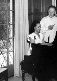 Plano medio de una mujer sentada al piano y Arthur Rubinstein observándola con un puro en la mano