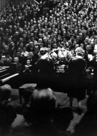 Plano general de Arthur Rubinstein (de perfil) y dos personas (de espaldas) entregándole una cesta de flores, con el público al fondo