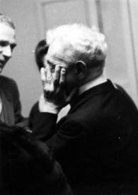 Plano medio de Arthur Rubinstein (perfil izquierdo) tapándose la cara con las manos y charlando con un hombre