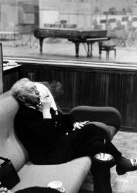 Plano general de Arthur Rubinstein (perfil derecho), sentado en un sillón, fumando un puro, escuchando las grabaciones