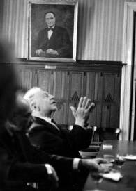 Plano medio de Arthur Rubinstein (perfil derecho) sentado en una mesa aplaudiendo con un retrato de un hombre al fondo.