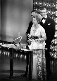 Plano general de Aniela Rubinstein hablando en el atril del escenario de la entrega de los Premios Óscar, con la estatuilla en la mano. Detrás François Reichenbach y Fred Astaire observandola