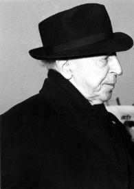 Plano medio de Arthur Rubinstein (perfil derecho) con un sombrero en un aeropuerto con el brazo izquierdo levantado