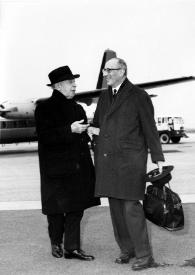 Plano general de Arthur Rubinstein (perfil derecho) charlando con un hombre (perfil izquierdo). Al fondo un avión.