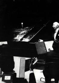 Plano medio de Arthur Rubinstein de pie, junto al piano, rodeado de la orquesta