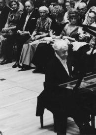 Plano medio de Arthur Rubinstein (perfil derecho) sentado al piano con el público detrás