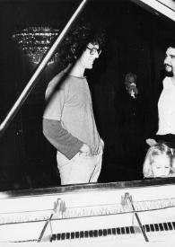 Plano medio de John Rubinstein (perfil derecho) observando a Jessica Rubinstein tocar el piano, detrás un hombre charla con John.