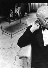 Plano general de Arthur Rubinstein (medio perfil derecho) sentado en un sillón tocándose la oreja derecha.