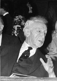 Plano medio de Arthur Rubinstein (medio perfil derecho) y Raymond Barre (Primer Ministro) (perfil izquierdo) hablando sentados entre el público.