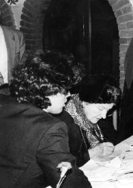 Plano medio de Aniela Rubinstein firmando un papel sentada en una mesa del restaurante, junto a ella en la misma mesa, un hombre, tres mujeres y un muchacho charlando