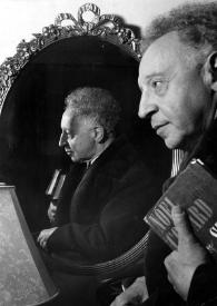 Plano medio de Arthur Rubinstein (perfil izquierdo) posando con un libro en la mano junto a un espejo donde también se ve su rostro