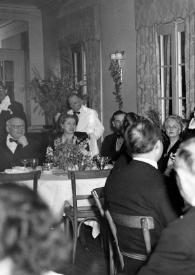 Plano medio de Arthur Rubinstein de pie entre las mesas con la copa alzada, hablando a los asistentes, entre ellos sentados en diferentes mesas: Sol Hurok