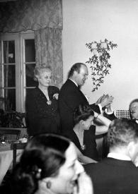 Plano general de una mujer y un hombre de pie saludando a Arthur Rubinstein sentado junto a una mujer y otros invitados