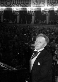 Plano medio de Arthur Rubinstein (perfil izquierdo) sentado al piano posando, al fondo el público