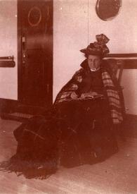 Plano general de la Señora de Rosentower sentada en una mecedora con una manta en las rodillas, posando en la cubierta del barco