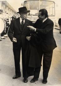 Plano general de Arthur Rubinstein y Serge Lifar charlando delante de un autobús, al fondo de la calle se observa una banda de músicos