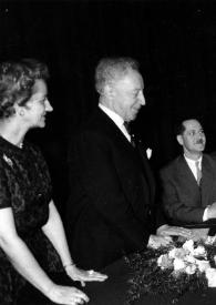 Portada:Plano medio de Aniela Rubinstein (perfil derecho) y Arthur Rubinstein (perfil derecho), en la mesa de la presidencia charlando con otras personas