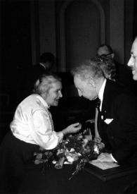 Portada:Plano medio de Arthur Rubinstein (perfil izquierdo), en la mesa de la presidencia, hablando con una mujer