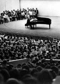 Portada:Plano general de Arthur Rubinstein (perfil derecho) sentado al piano en el escenario jutno al público. Fotografía tomada desde el fondo de la sala