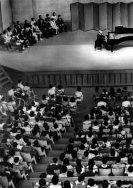 Portada:Plano general de Arthur Rubinstein (perfil derecho) sentado al piano mientras el público le escucha. Fotografía tomada desde el fondo de la sala de conciertos.