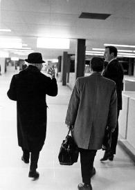 Portada:Plano general de Arthur Rubinstein (de espaldas) caminando y charlando por el aeropuerto con Louis Bender y un hombre del aeropuerto