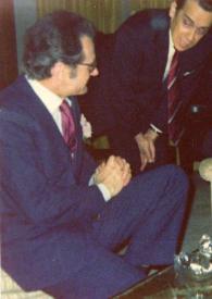 Portada:Plano general de dos hombres, uno de pie y otro sentado, charlando con Arthur Rubinstein (perfil izquierdo) fumando un puro, sentado en un sillón