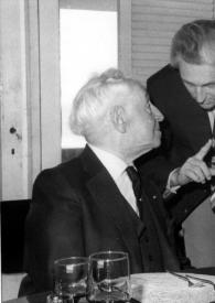 Portada:Plano medio de Arthur Rubinstein sentado en una mesa hablando con Shlomo Lahat de pie, junto a ellos la Señora de Freda Cocks sentada en la mesa escuchándoles.