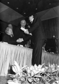 Portada:Plano medio de Aniela Rubinstein, Jan Jacob Bistritzky y un hombre sentados en la mesa presidencial, Aniela le entrega un diploma a Benjamin Frith