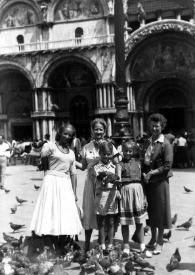 Portada:Plano general de Eva, Aniela, John y Alina Rubinstein posando junto a una mujer dando de comer a las palomas en la Plaza de San Marco