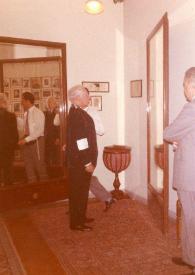 Portada:Plano general de Arthur Rubinstein (perfil derecho) probándose un traje frente a un espejo, el sastre detrás