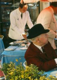 Portada:Plano medio de Arthur Rubinstein (perfil derecho) con sombrero, sentado en la mesa de la terraza de un restaurante con otros hombres, hablando con el camarero